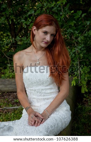 Pretty redhead bride in her wedding dress, sitting outside on a log in a grassy field, looking at the camera with a thoughtful expression