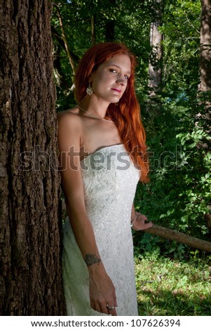 Pretty redhead bride in her wedding dress, standing outside by a tree and  looking at the camera with a thoughtful expression