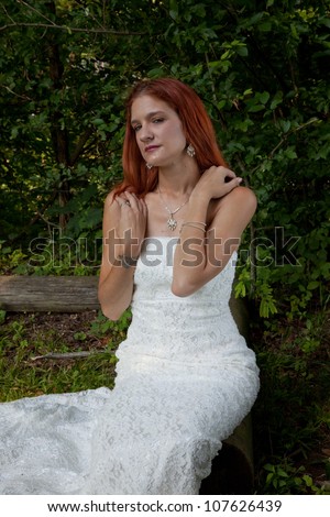 Pretty redhead bride in her wedding dress, sitting outside on a log in a grassy field, looking at the camera with a thoughtful expression