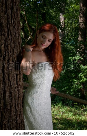 Pretty redhead bride in her wedding dress, standing outside by a tree and  looking at the camera with a thoughtful expression