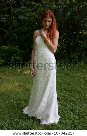 Lovely redhead woman in a white wedding dress,standing in a field of green grass with woods in the background