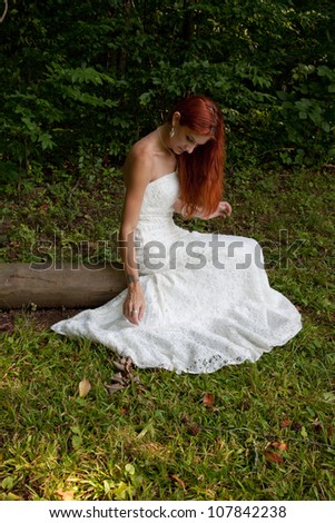 Lovely redhead woman in a white wedding dress, sitting in a field of green grass with woods in the background