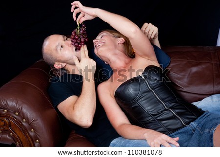 Couple reclining on a couch together, she is feeding him grapes by dangling them for him to bite