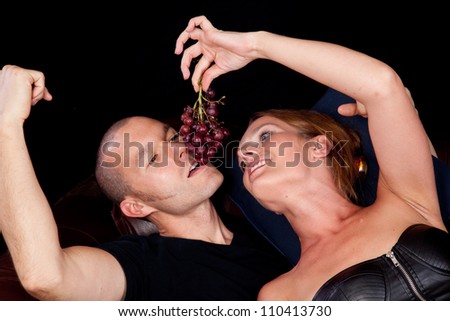 Couple reclining on a couch together, she is feeding him grapes by dangling them for him to bite