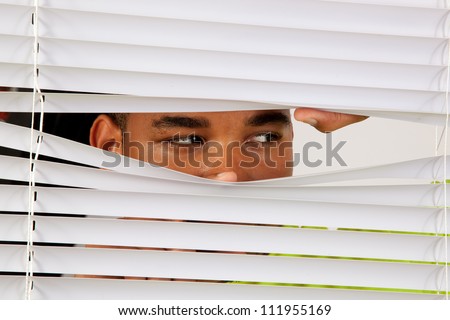 Black man behind window blinds and peering out like he was watching someone or spying