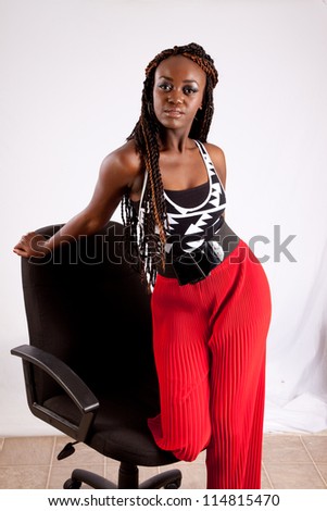 Lovely black woman in red slacks kneeling in a black business chair with one hand on  the back of the chair and looking at the camera with a thoughtful, friendly expression