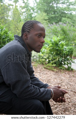 Thoughtful, pensive African American man sitting thoughtfully outside in nature, with trees in the background