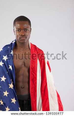 Serious black man with no shirt on, showing his muscular torso, with an American flag draped over his shoulders and an intense expression for the camera