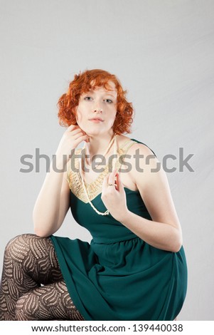Pretty redhead woman in a green dress, sitting on a stool and looking at camera with a  thoughtful, friendly expression with her chin on her hand and playing with her pearls