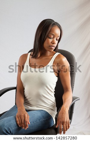 Lovely black woman in a white, tank top, sitting and holding her left arm with her right hand, while she looks down thoughtfully