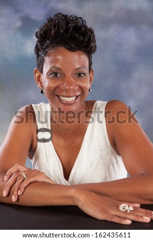 Pretty black woman sitting in a white dress, her arms crossed on a table, and looking at the camera with a friendly, happy smile