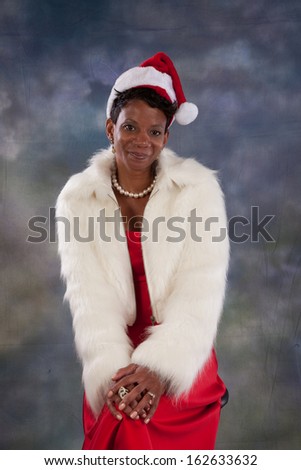 Pretty black woman wearing a red dress and white fur jacket, and a red and white Santa Claus hat, looking at the camera with a happy smile