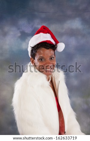 Pretty black woman wearing a red dress and white fur jacket, and a red and white Santa Claus hat, looking at the camera with a happy smile