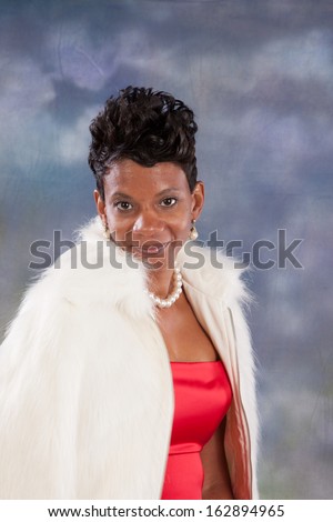 Pretty black woman  in a red sleeveless dress and white fur jacket, looking  at the camera  with a bright, happy smile