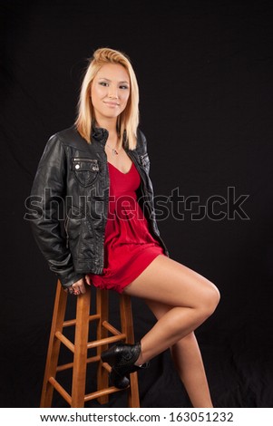 Pretty blond woman in black leather jacket and short red dress, sitting on a wooden stool and  looking at the camera with a serious, thoughtful expression