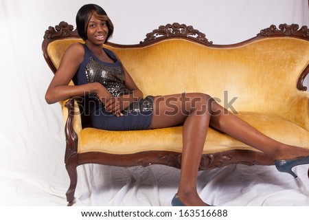 Black woman in a black dress, sitting on a couch and looking at the camera with a happy, friendly smile