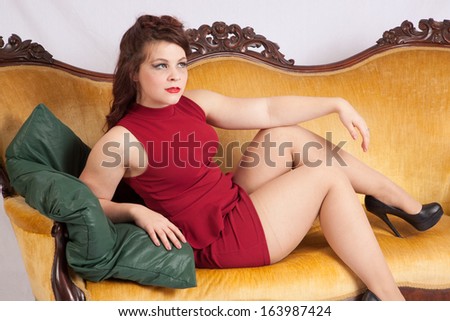 Pretty Caucasian woman in a short red dress, sitting on an old couch and looking with a relaxed flirting expression