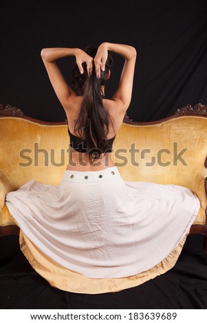 Pretty woman in black top and dress sitting  on a gold couch with her back to the camera and playing with her hair