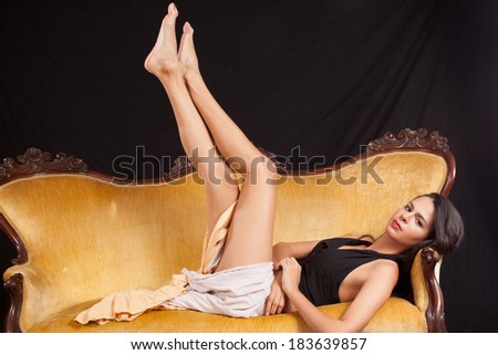 Pretty woman in black top and dress laying back  on a gold couch with her legs raised up in the air, looking at the camera with a serious expression