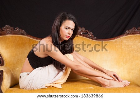 Pretty woman in black top and dress, sitting on a gold couch with her feet pulled up on the couch and looking at the camera with a serious expression