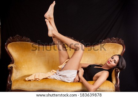 Pretty woman in black top and dress laying back  on a gold couch with her legs raised up in the air, looking at the camera with a serious expression