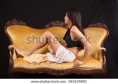 Pretty woman in black top and dress sitting  on a gold couch with her knees bent and feet on the couch