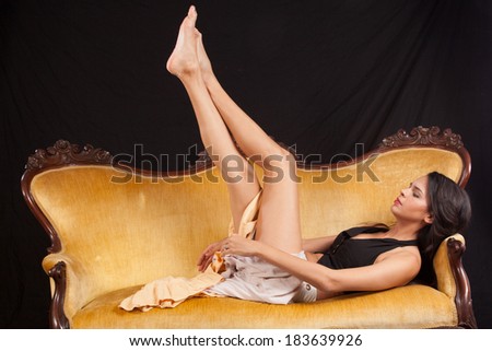 Pretty woman in black top and dress laying back  on a gold couch with her legs raised up in the air,