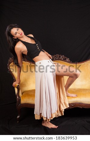 Pretty woman in black top and dress, standing with her foot on a couch and looking at the camera