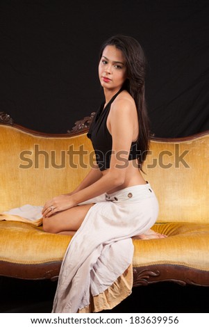 Pretty woman in black top and dress sitting on a gold couch with her knees bent and feet under her, and looking at the camera with a serious expression