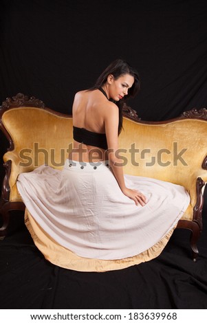 Pretty woman in black top and dress sitting  on a gold couch with her back to the camera