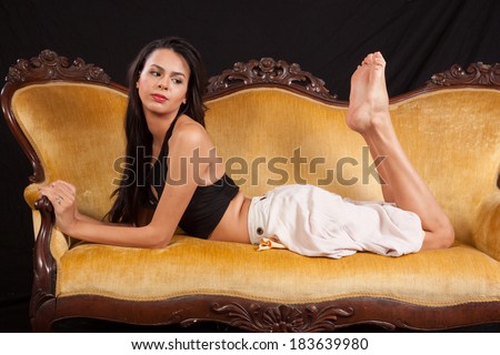 Pretty woman in black top and dress,reclining on a gold couch with her knees bent and feet in the air, and looking at the camera with a serious expression