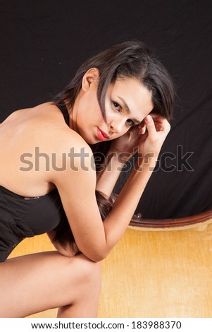 Pretty woman in black top  standing by a gold couch with one foot on the couch and leaning forward,