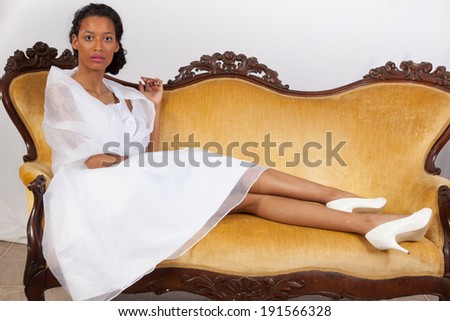 Lovely black woman in white dress, sitting on a gold couch, looking at the camera with a friendly, thoughtful expression