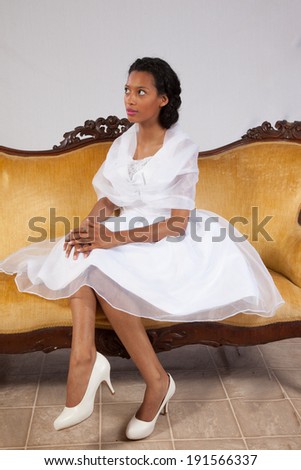 Lovely black woman in white dress, sitting on a gold couch, looking left with a friendly, thoughtful expression