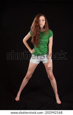 Pretty woman in green blouse and white shorts and barefoot, with long hair and one hand on her hip  looking at the camera thoughtfully