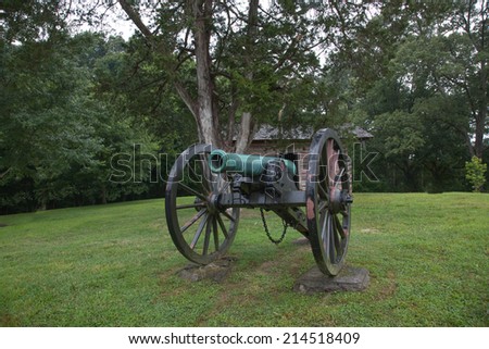 Old canon sitting before a log cabin in a field, with trees in the background