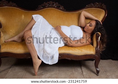 Pretty pregnant woman laying down on a gold couch and holding her belly with a happy expression.