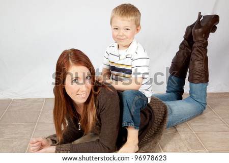 A mother and her young son playing together as he sits on her back as she lays on the floor  and they laugh together