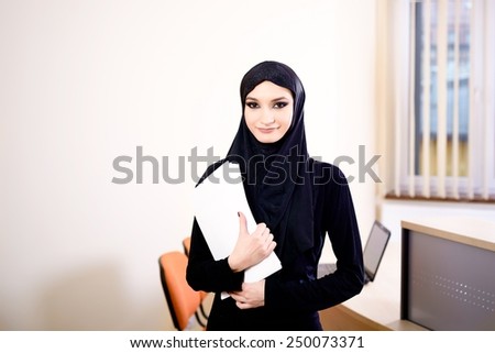 Woman dressed in black and wearing the hijab while working in the office.  In her hand she holds the documents