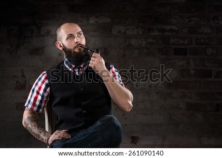 Thoughtful bearded man in a checkered shirt smoking a pipe sitting in front of a brick wall