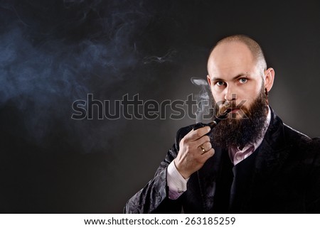 bald bearded man wearing old-fashioned suit and smoking a pipe. A lot of smoke in the picture. Photo taken in the studio on a gray background