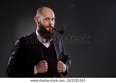 Bald bearded man holding a smoking pipe in his mouth. He is dressed in a black jacket. Photo taken in the studio on a gray background
