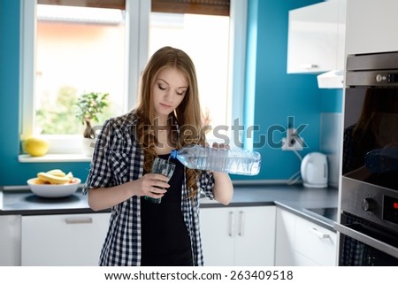 Young blonde woman in kitchen woman pouring a glass of water. Standing in a bright modern kitchen. Dressed in a plaid shirt with short sleeve