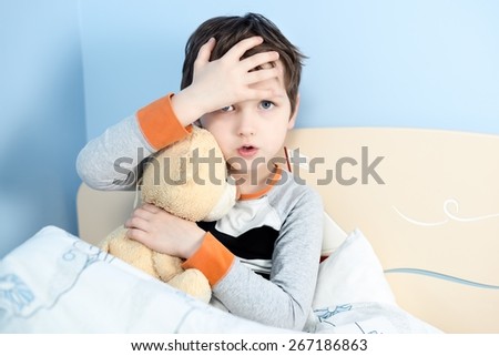 Sick little boy hugs his teddy bear in bed. Touching his forehead to check temperature