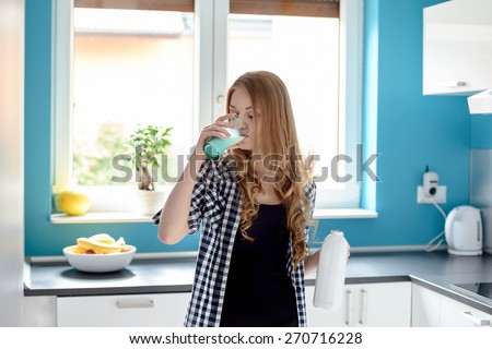 Thirsty young blond woman drinking milk from a glass in the kitchen. Holding the bottle in the other hand