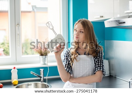 Beautiful young blond woman standing in front of the kitchen window, holding in her hand mixer