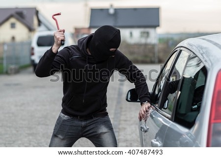 Man dressed in black with a balaclava on his head breaking a glass in car with crowbar. Car thief, car theft concept