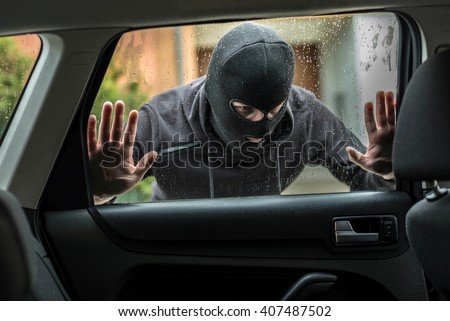 Man dressed in black with a balaclava on his head looking through car window and wondering how to break into this car. Car thief, car theft concept