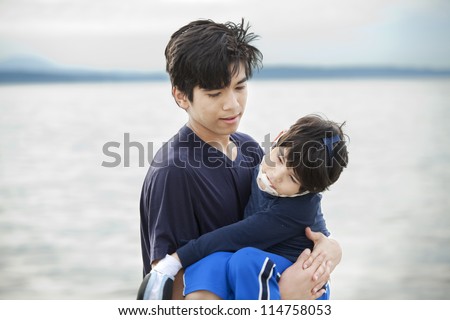 Big brother carrying disabled boy on beach by water. Child has cerebral palsy