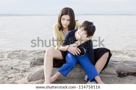 Tired teenage sister holding her disabled brother on the beach. Child has cerebral palsy